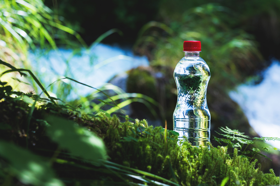 Transparent plastic A bottle of clean water with a red lid stands in the grass and moss on the background of a rugged mountain river. The concept of pure natural drinking water.