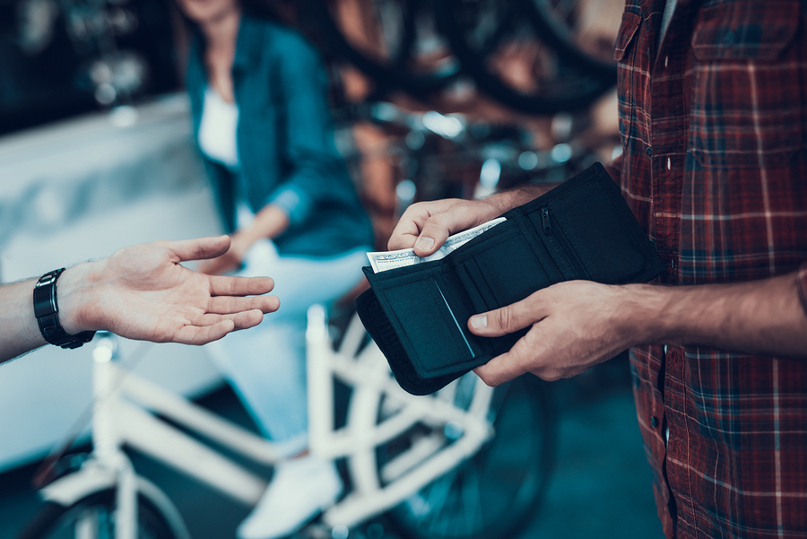 Closeup of Male Hand Gives Money From Purse to Seller. Shop Assistant Hand Takes Cash with Blurred Customers and Bikes on Backround in Sport Bicycle Store. Cash Payments Concept