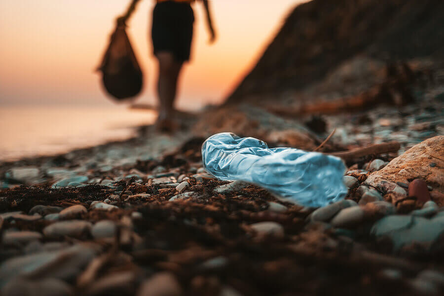 Save Download Preview Close up of dirty blue plastic bottle lying on the beach. Defocused silhouette of a person holding a plastic bag with garbage on the background.
