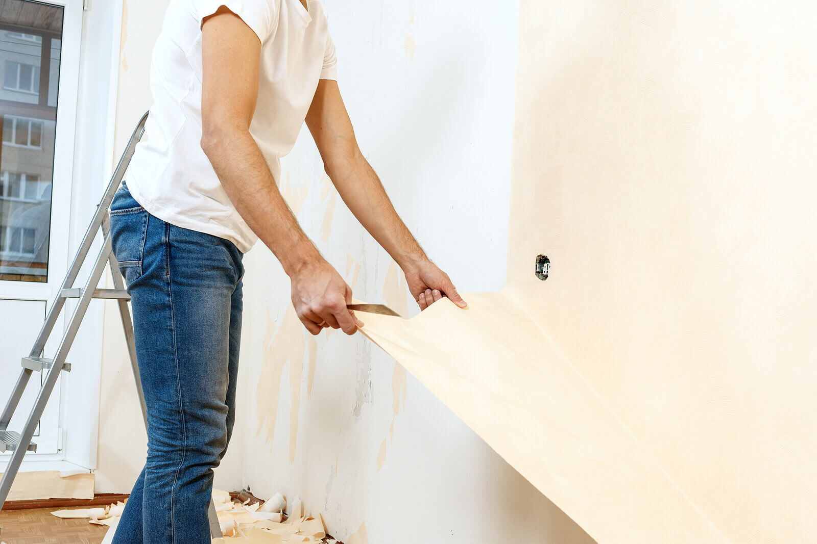 Side view of a man removing wallpaper from a wall, with a set of ladders in the background.